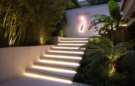 Night time in a london garden with floating steps lit by LED strip lights. Steps lead to neon sculpture of a nail head on a rendered wall. Surrounded by lush tropical plants lit by garden spot lights.