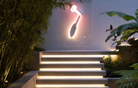 Night time in a london garden with floating steps lit by LED strip lights. Steps lead to neon sculpture by James Clar of a nail head on a rendered wall. Surrounded by lush tropical plants lit by garden spot lights.