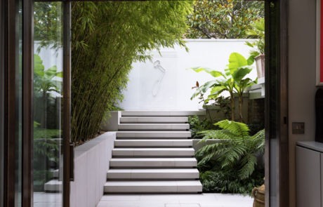 Open doors from basement looking to sleek contemporary garden. Porcelain paving and floating steps with lush tropical planting.